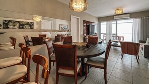 A spacious dining area with a large round table and multiple chairs, a chandelier, and a television in the background. - CONDOMINIUM CLUB (RESORT) RIVIERA 4 ROOMS - ALL TIME (Bertioga)