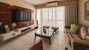 A living room with a flat-screen TV mounted on a wooden panel, a glass coffee table, a sofa, and a chair. - CONDOMINIUM CLUB (RESORT) RIVIERA 4 ROOMS - ALL TIME (Bertioga)