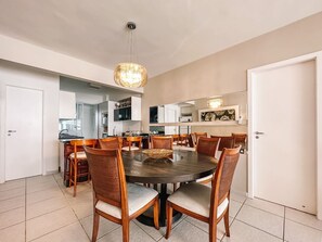 Uma sala de jantar com uma mesa redonda cercada por cadeiras, uma cozinha ao fundo e uma porta à direita.