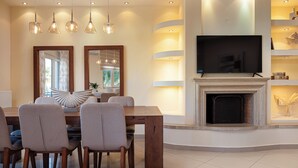 Uma sala de estar moderna com lareira, televisão montada acima e uma área de jantar com cadeiras e mesa.