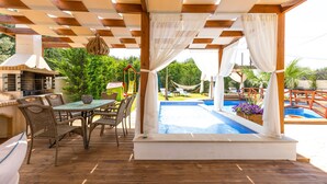 Um pátio externo com piscina, deck de madeira, móveis de vime e área de churrasco.