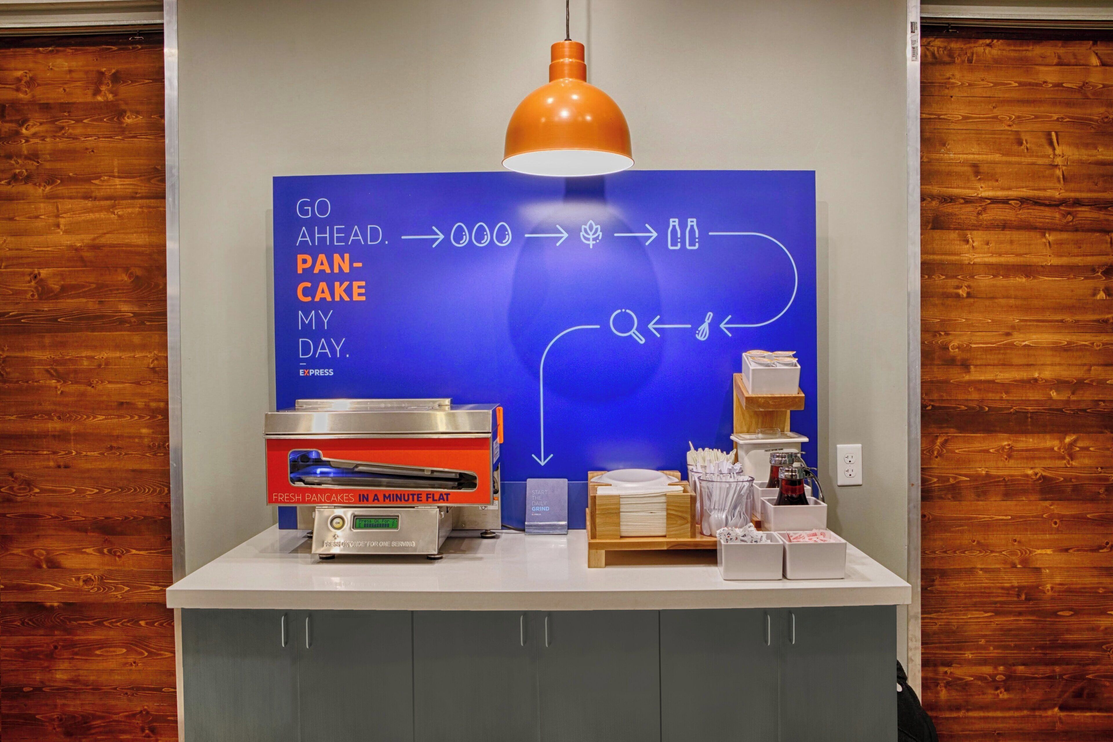 A kitchen counter with a flat-screen TV displaying a breakfast menu, a coffee machine, and a sign for fresh pancakes.