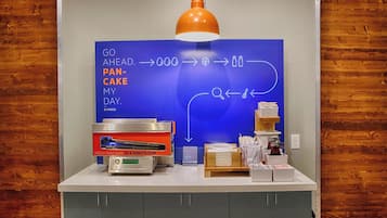 A kitchen counter with a flat-screen TV displaying a breakfast menu, a coffee machine, and a sign for fresh pancakes.