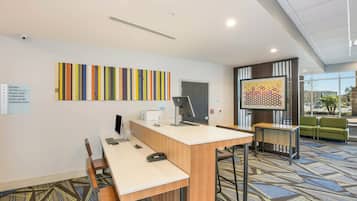 Un espace d’accueil d’hôtel comprenant un comptoir de réception, un ordinateur et un coin salon. Des panneaux indiquent l’emplacement des chambres et du centre d’affaires.