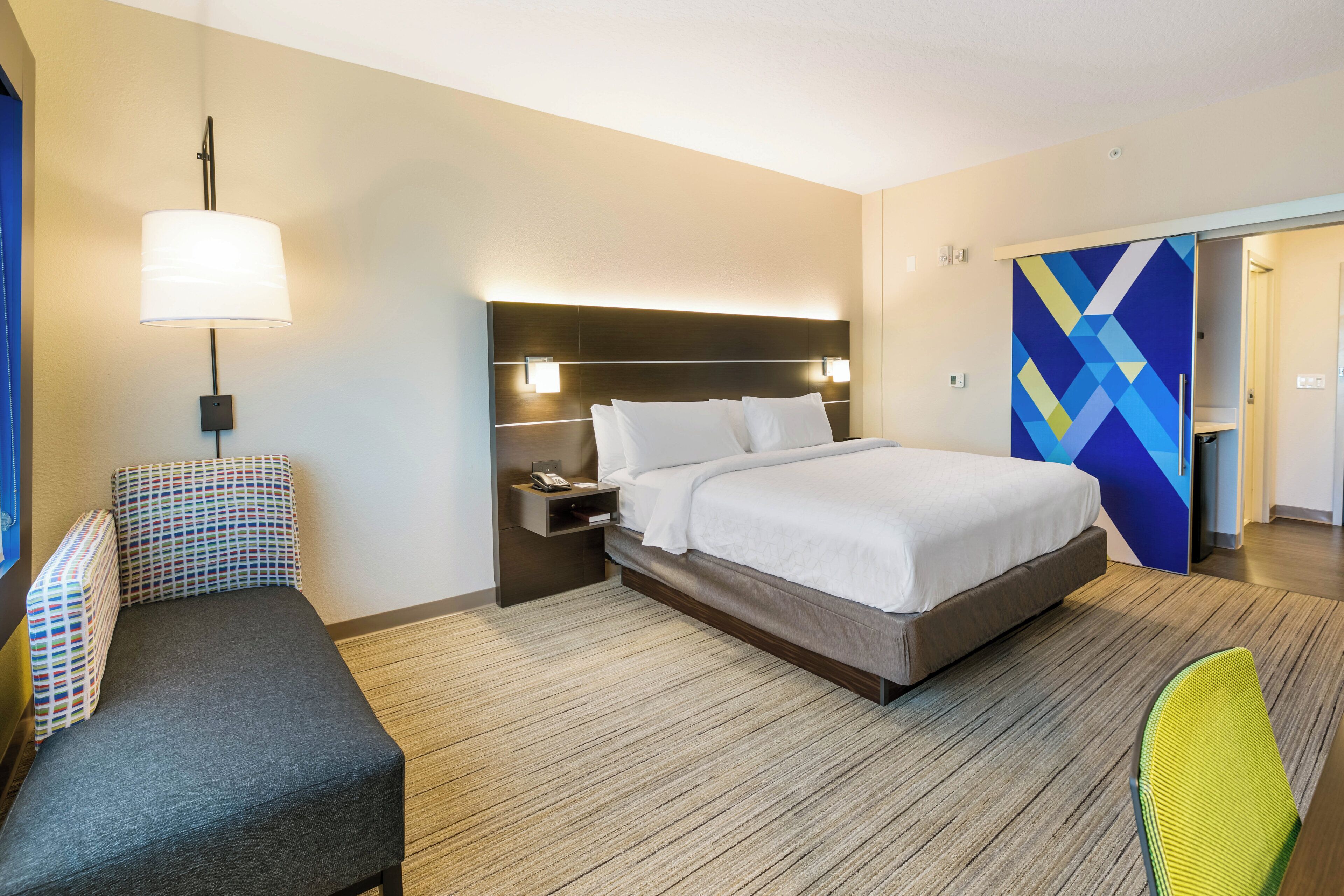 A hotel room with a bed, bedside table, lamp, chair, and a colorful wall decoration.
