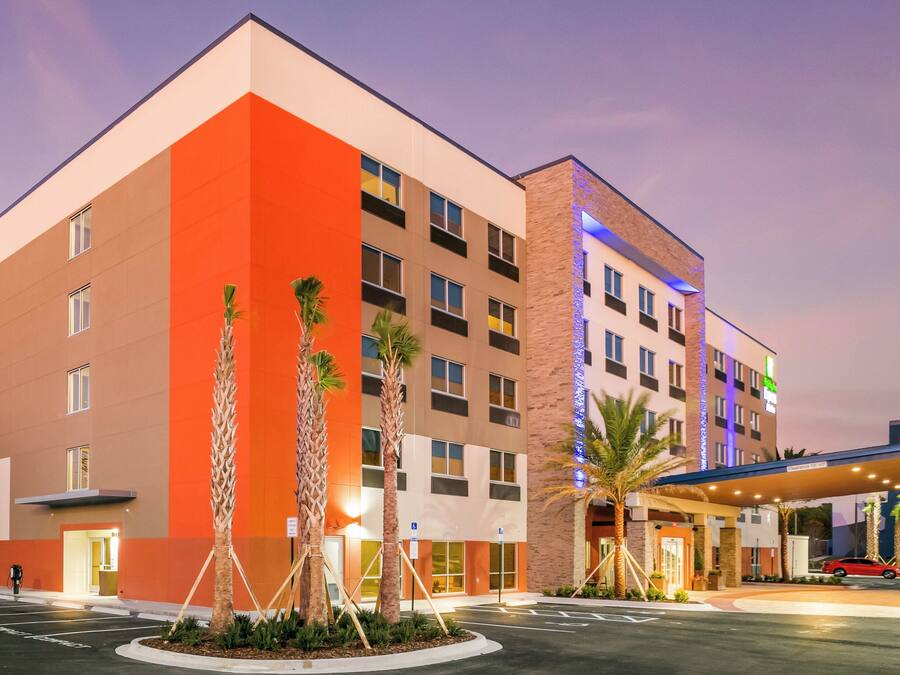 A multi-story hotel with a modern design, featuring a prominent orange facade and a parking lot with palm trees.