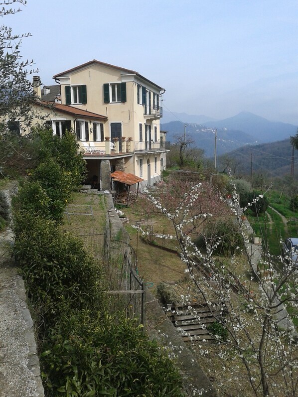A two-story building with a balcony, surrounded by greenery and a mountainous backdrop.