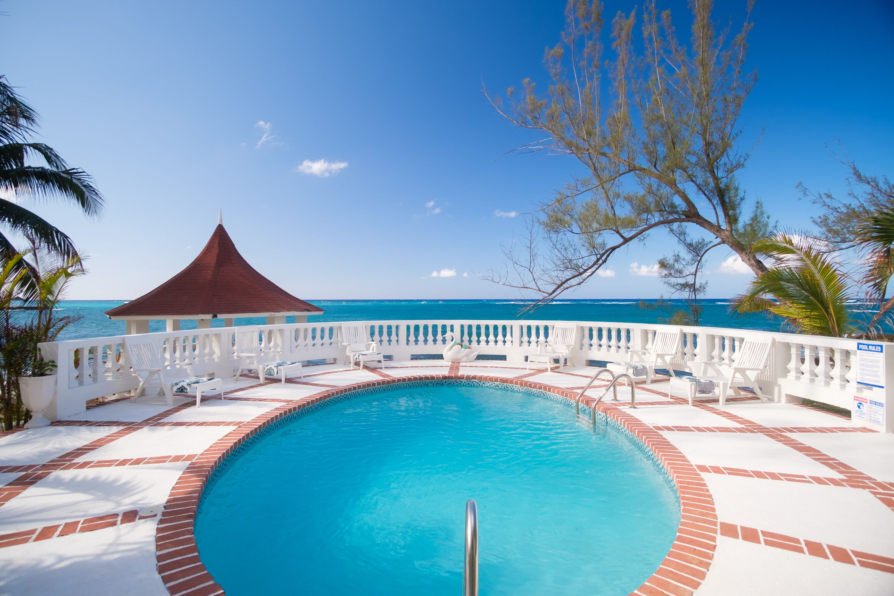 A swimming pool with a red-roofed pavilion, white lounge chairs, and a view of the ocean.