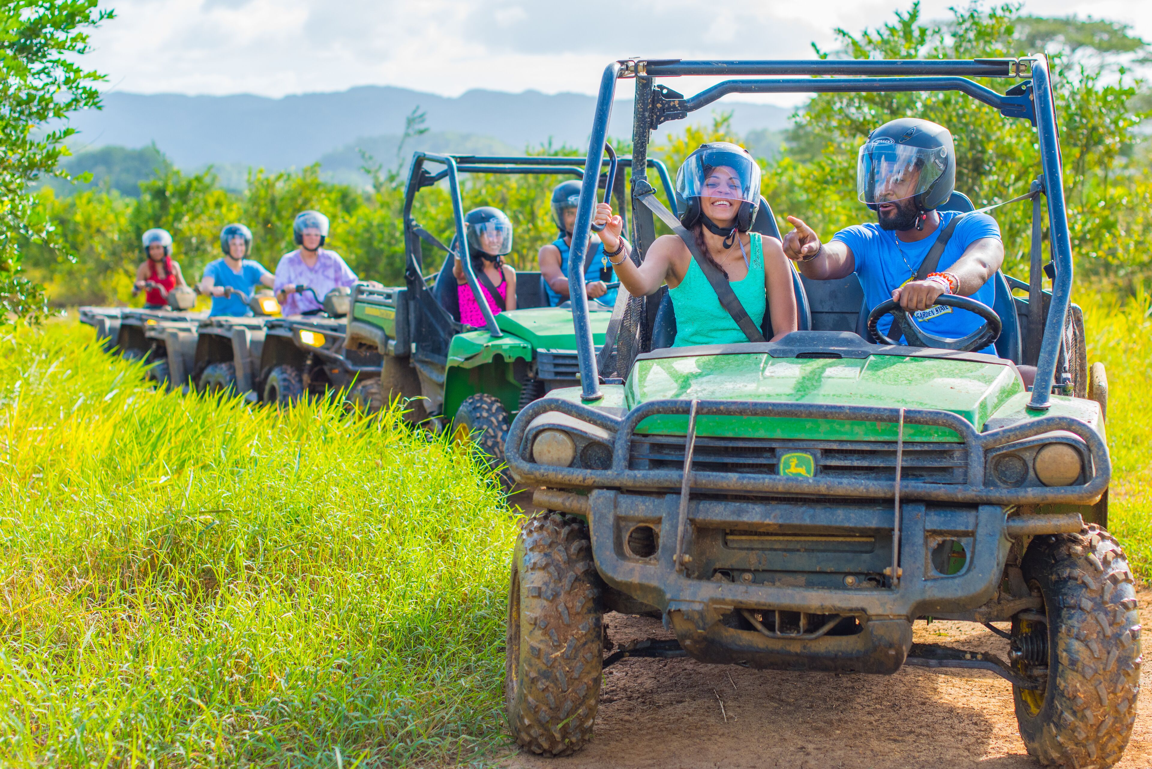 A green off-road vehicle with passengers wearing helmets, driving on a dirt path surrounded by greenery.
