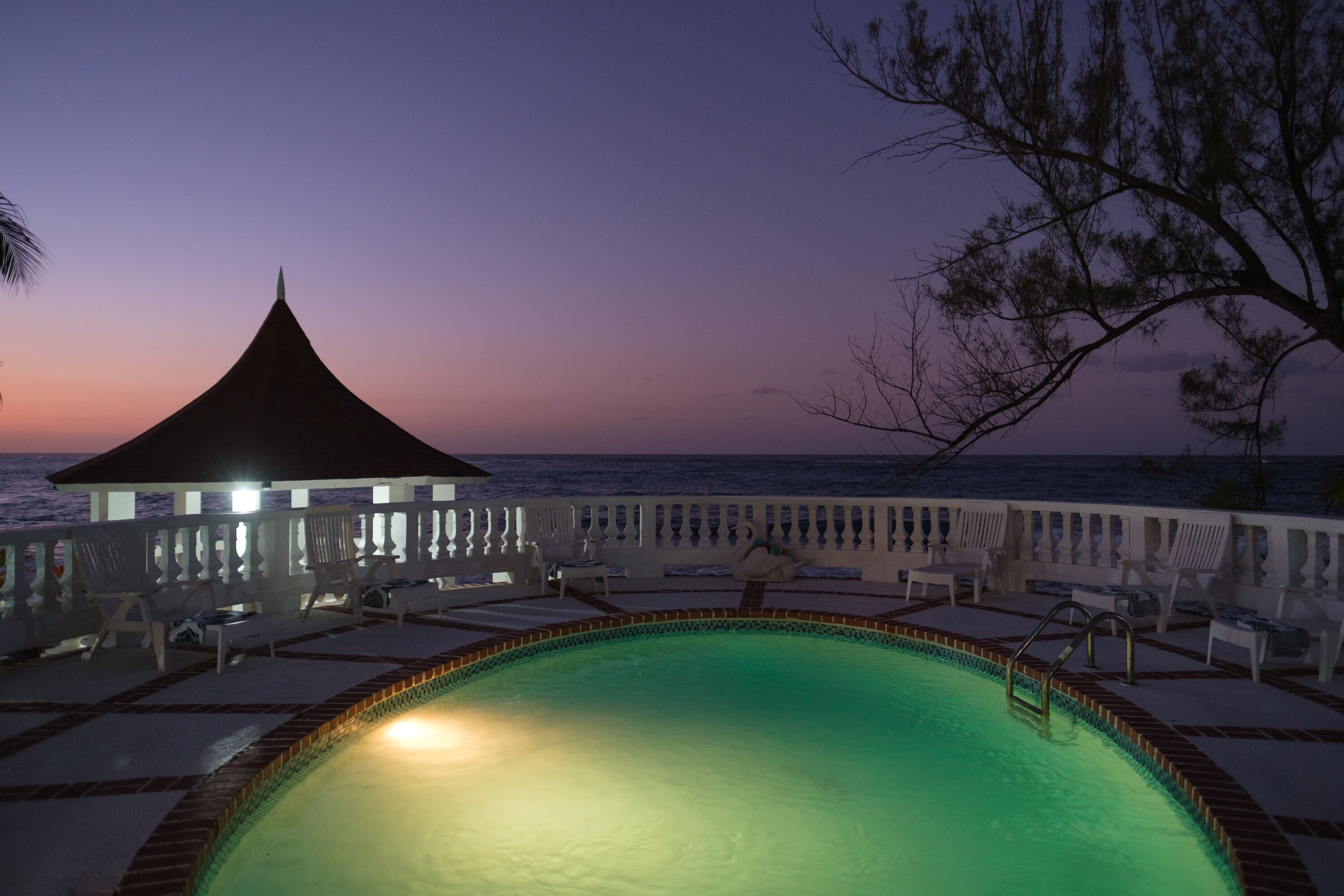 A pool area with lounge chairs and a gazebo overlooking the sea at dusk.