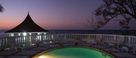 A pool area with lounge chairs and a gazebo overlooking the sea at dusk.