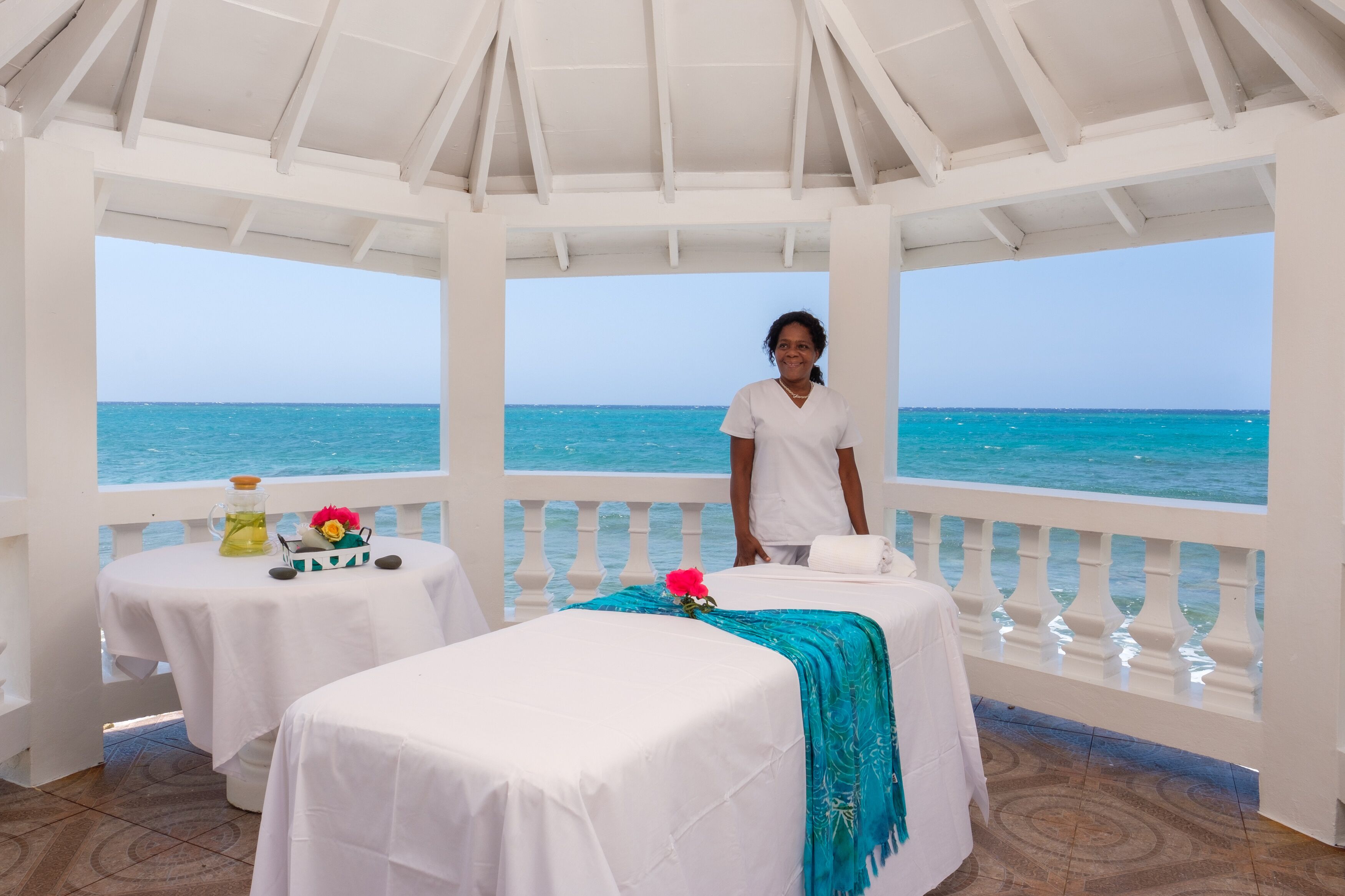 A serene spa area with a view of the ocean, featuring two massage tables and a small table with a bottle and flowers.