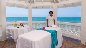 Een rustgevende spa-ruimte met uitzicht op de oceaan, met twee massagetafels en een kleintje met een drankje en bloemen.