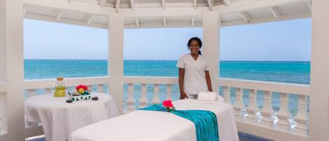 A serene spa area with a view of the ocean, featuring two massage tables and a small table with a bottle and flowers.