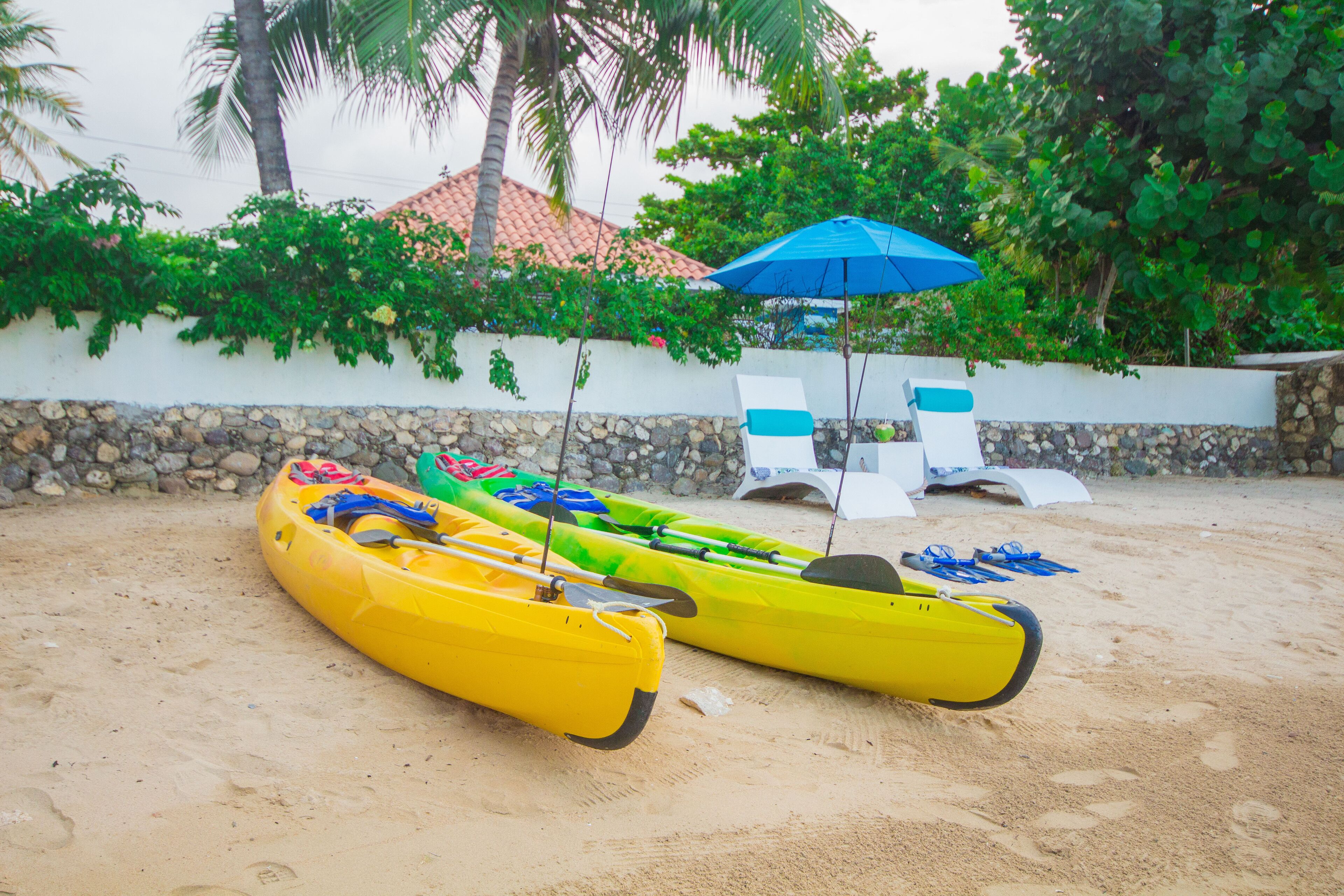 Deux kayaks jaunes sur une plage de sable, avec un mur blanc et un parasol bleu en arrière-plan.