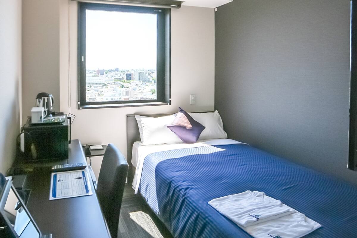 A hotel room with a bed, a desk, a chair, a window with a city view, and a coffee maker.