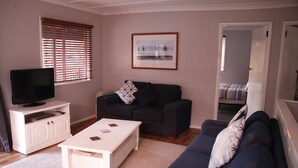 Un salon avec un téléviseur posé sur un meuble blanc, un canapé foncé avec un coussin, une table basse blanche et un tableau encadré accroché au mur.