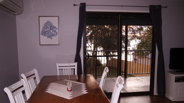 Un coin repas avec une table en bois, des chaises blanches, une œuvre d’art encadrée et une vue sur l’extérieur, accessible par une porte-fenêtre coulissante, donnant sur un espace extérieur avec une clôture et des arbres.