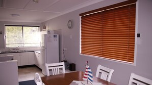 Un coin repas avec une table en bois, des chaises blanches, une décoration sur le thème d’un voilier, une fenêtre avec des stores et, au fond, une cuisine.