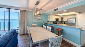 Een eetgedeelte met een tafel en stoelen, een keuken met moderne apparatuur, en uitzicht op de oceaan door grote ramen.