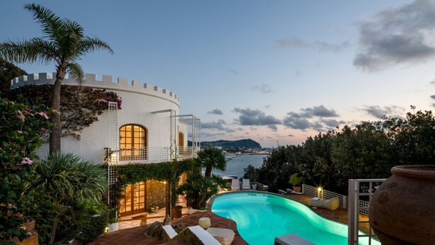 Un edificio blanco con una torre redonda, piscina y vistas al mar.