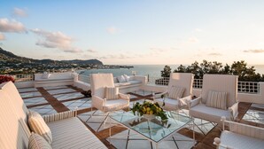Una terraza con mobiliario blanco, una mesa de vidrio y vista al mar y a una ciudad distante.