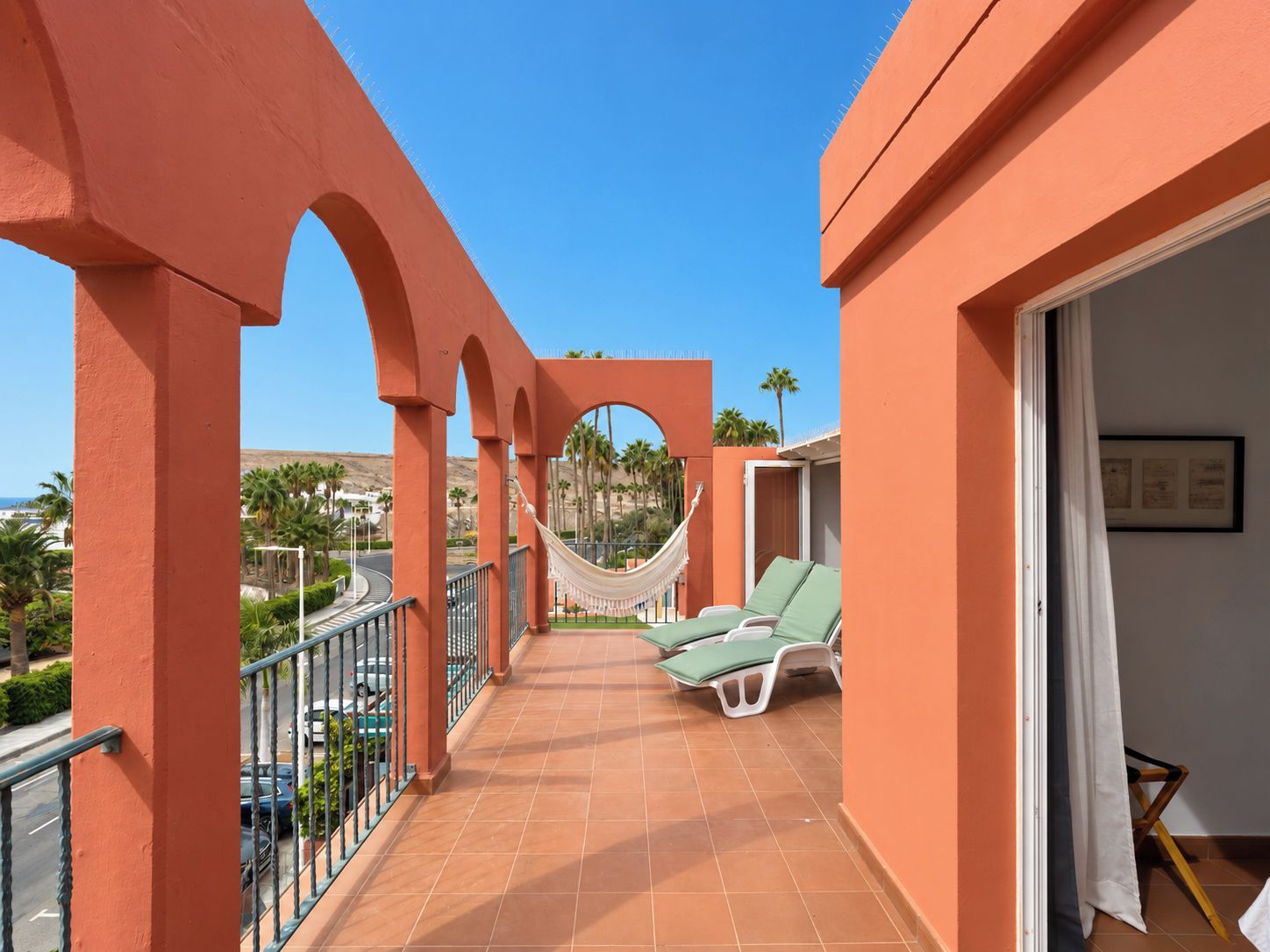 A balcony with a view of palm trees and a road, a hammock, and two sun loungers.