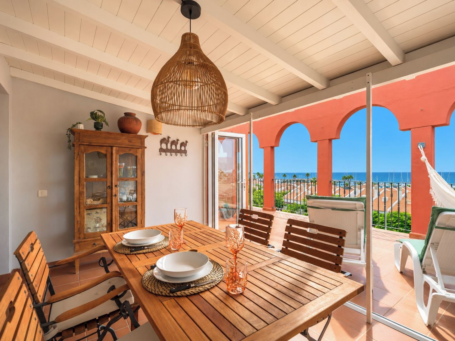 Una mesa de comedor de madera dispuesta con platos y vasos, una lámpara colgante y vista a la playa a través de un ventanal.