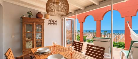 A wooden dining table set with plates and glasses, a hanging light fixture, and a view of the beach through a glass door.