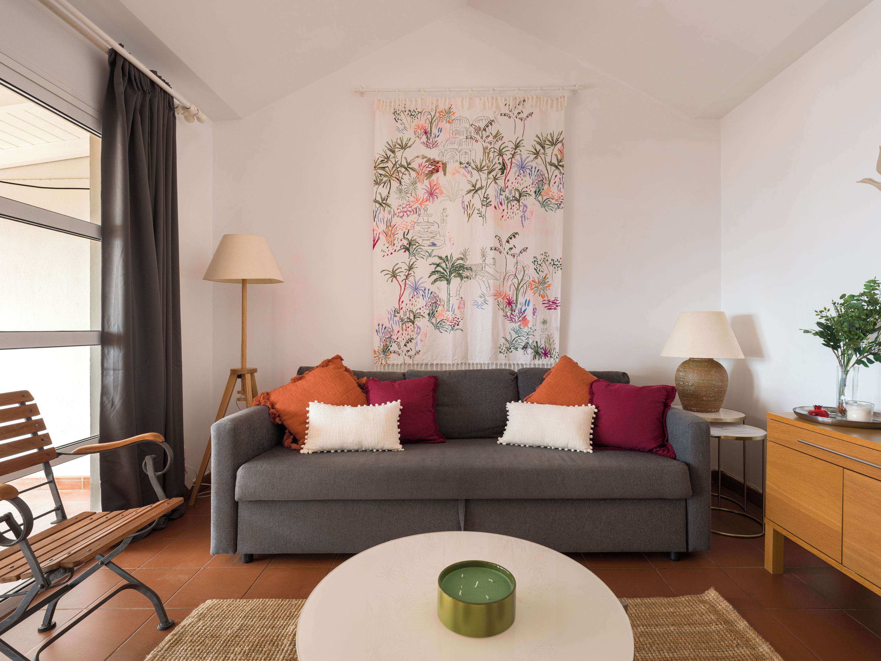 Un salon avec un canapé gris, une table basse ronde, une lampe, un banc et une fenêtre avec des rideaux.