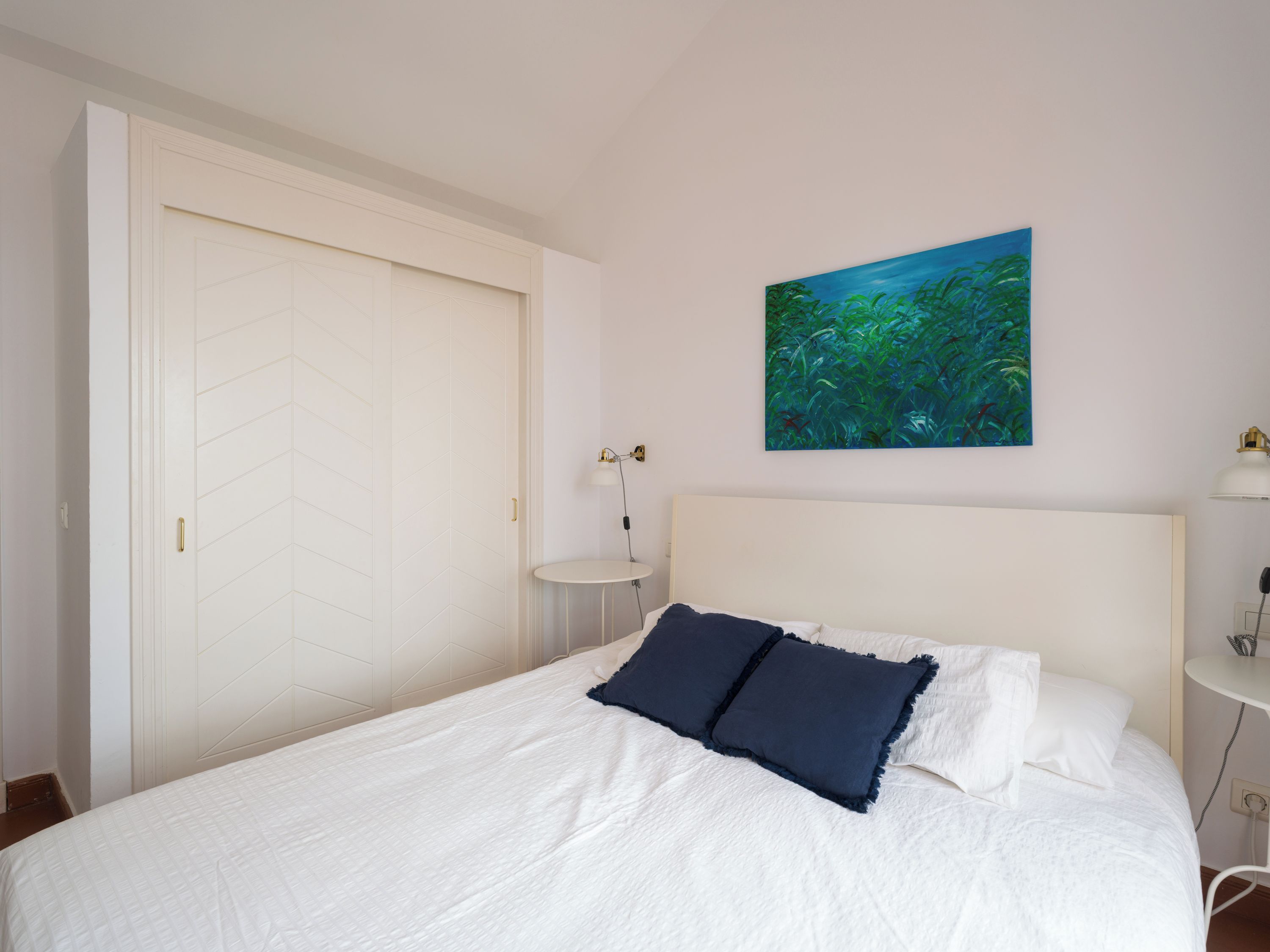 A bedroom with a bed, a white door, a nightstand, a lamp, and a painting on the wall.