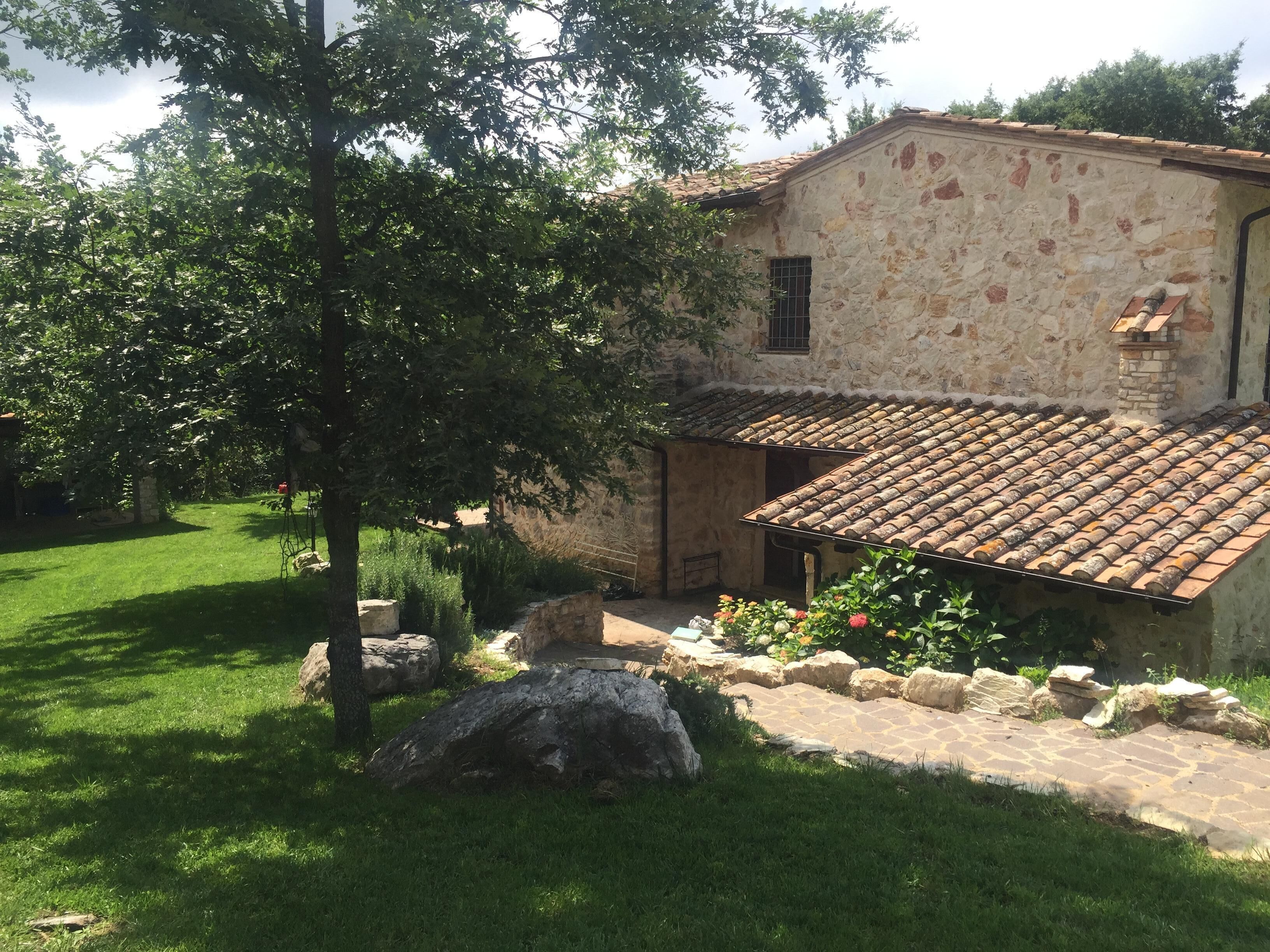 Un bâtiment en pierre avec un toit en tuiles, entouré d’une pelouse bien entretenue et d’un grand arbre.