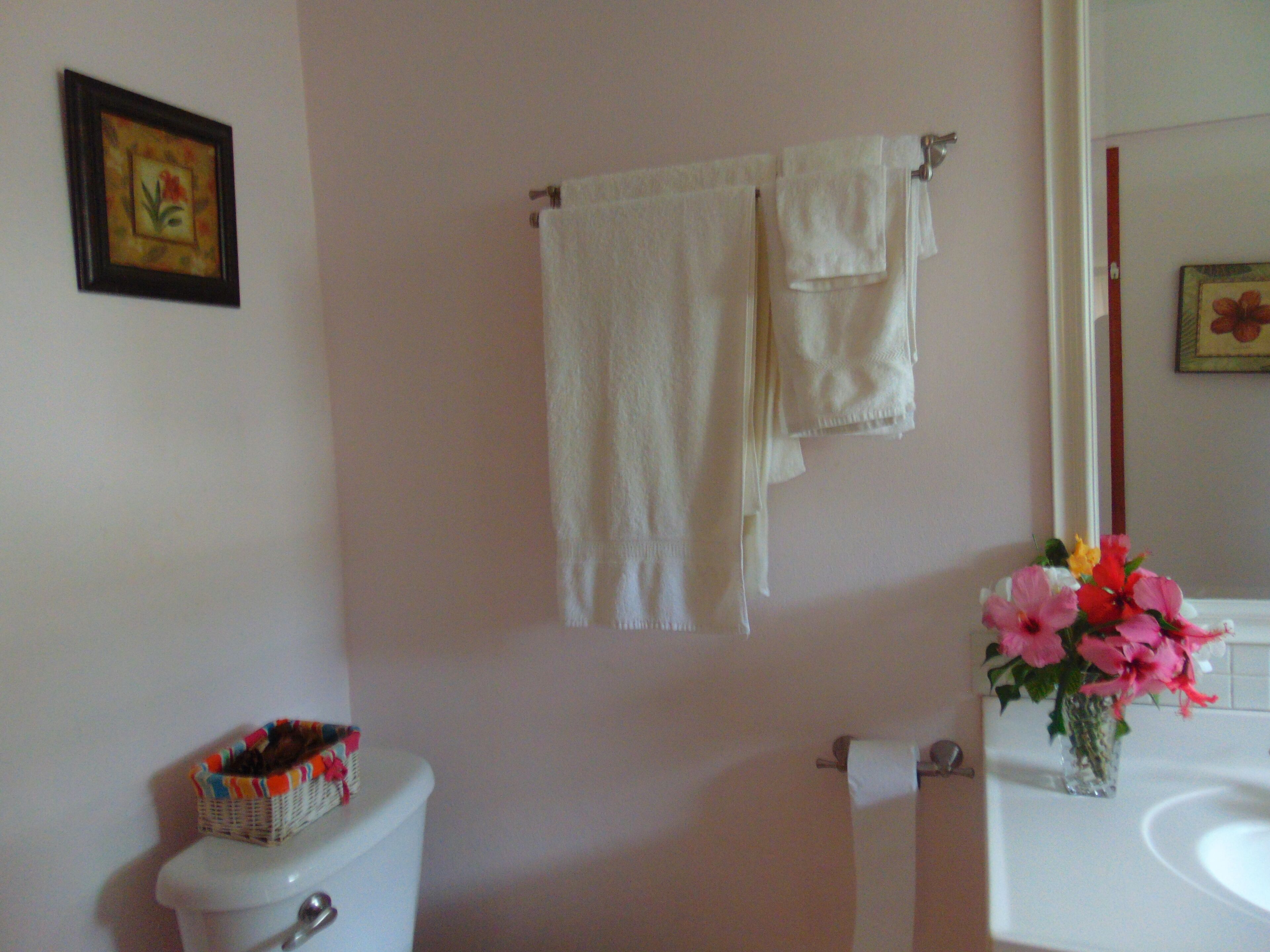 A bathroom with a toilet, a sink, and a towel rack with two towels. There is a basket on the toilet and a vase with flowers on the sink.