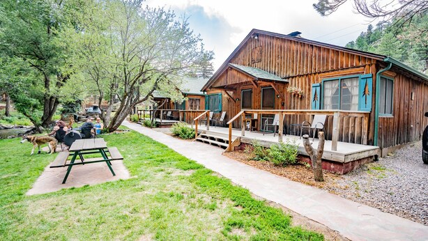 Un chalet en bois rustique, doté d’un porche, entouré de verdure et bordé d’un petit ruisseau.