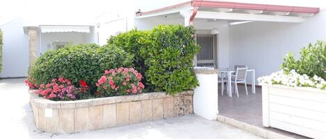 Ein überdachter Patio-Bereich mit weißem Tisch und Stühlen, umgeben von einer niedrigen Steinmauer mit verschiedenen Pflanzen und Blumen.
