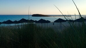 A beach with thatched umbrellas and tall grass in the foreground, a rocky outcrop in the middle distance, and calm sea under a clear sky.