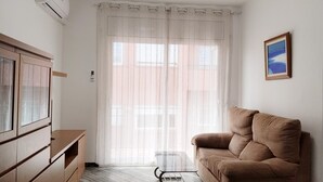 A room with a sofa, a glass coffee table, a cabinet, a window with sheer curtains, and a wall-mounted air conditioner.