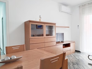 Une pièce comprenant une table à manger en bois, des chaises, un meuble avec des portes en verre, un téléviseur et un appareil de climatisation.