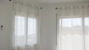 A hotel room with a bed, two windows with curtains, a towel on the bed, and a view of buildings outside.