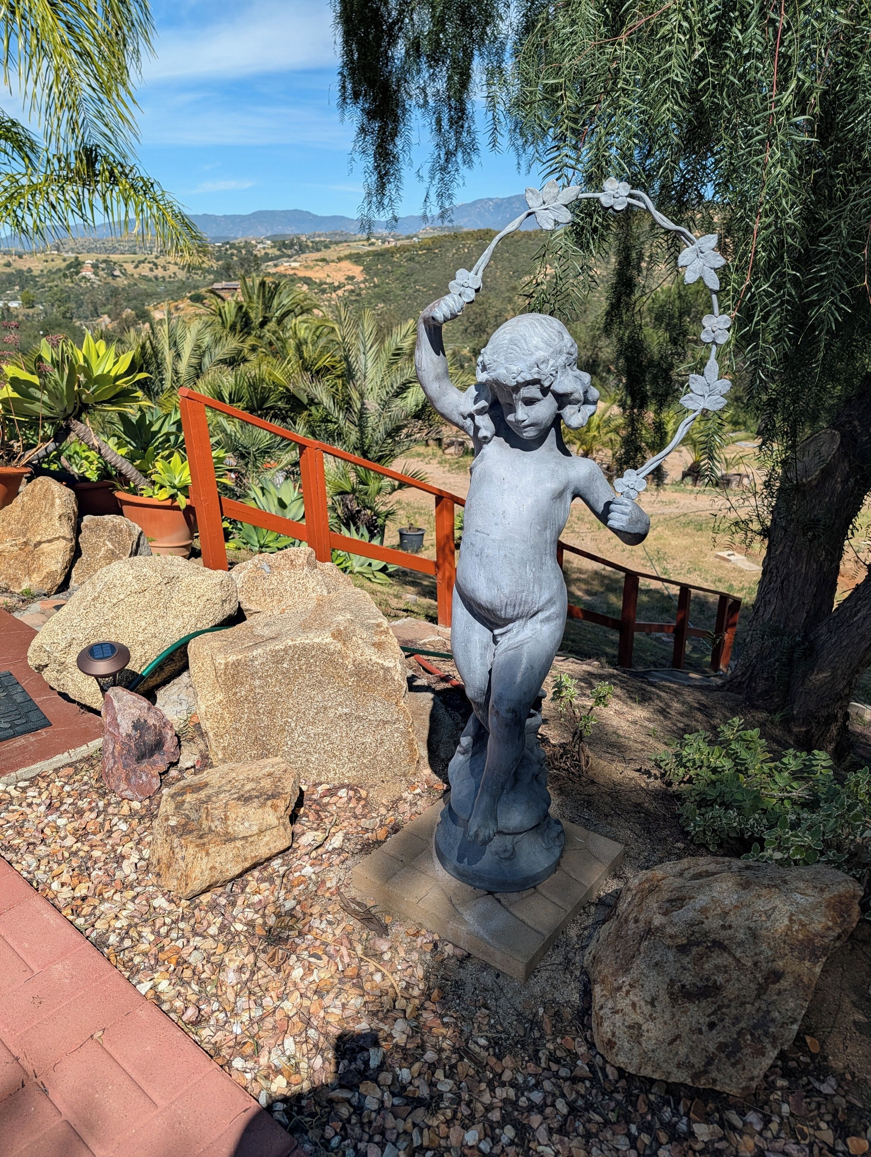 A statue of a child holding a flower, surrounded by rocks and plants, with a wooden fence and a tree in the background.