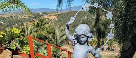 A statue of a child holding a flower, surrounded by rocks and plants, with a wooden fence and a tree in the background.