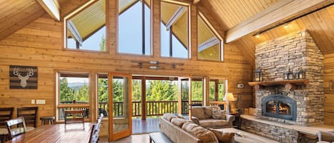 A spacious loft-style room with a high ceiling, large windows, a stone fireplace, and a wooden dining area.