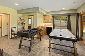 A recreational room with a foosball table, a shuffleboard table, and a kitchen area with wooden cabinets.