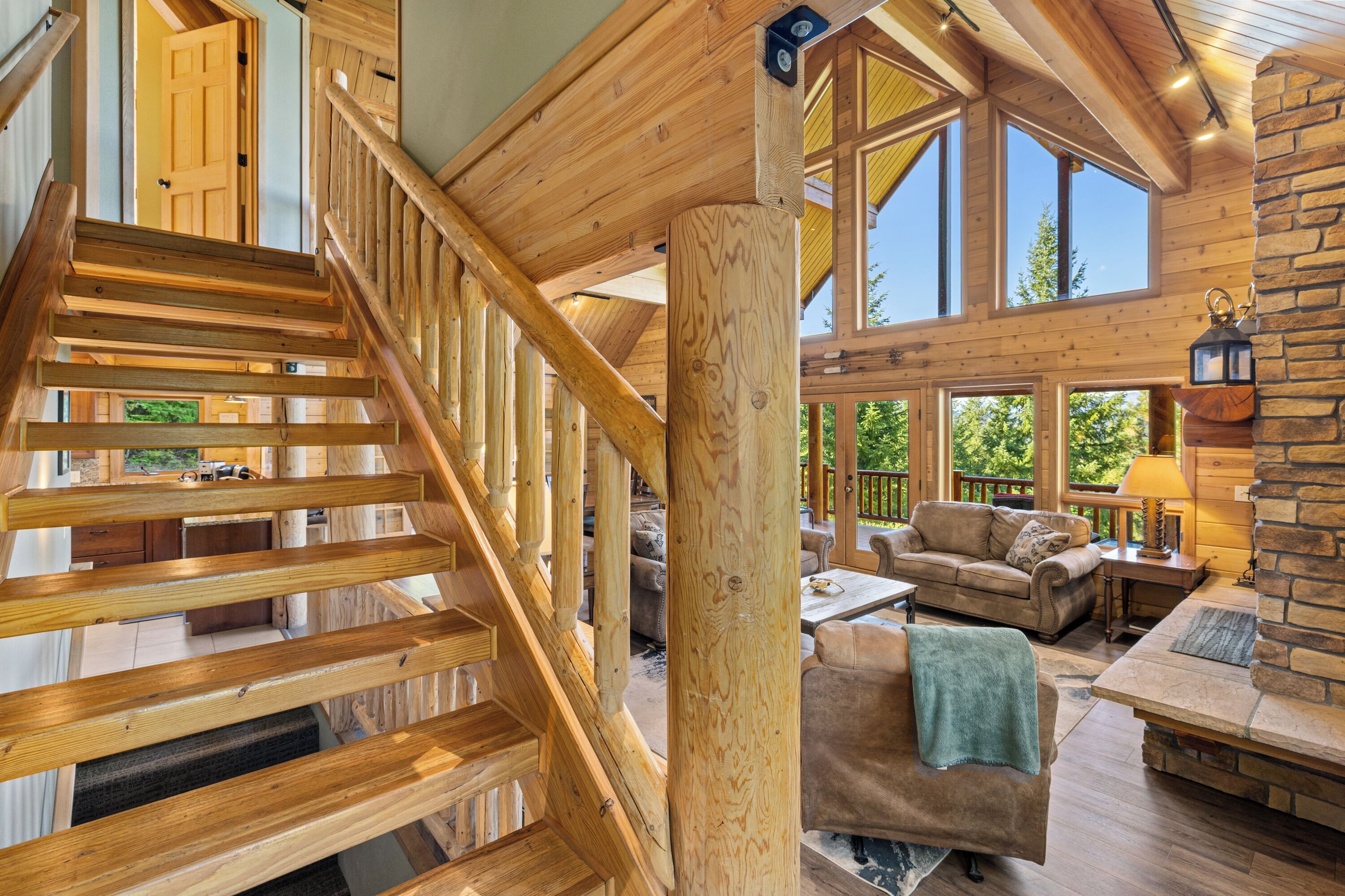 Un escalier en bois mène à une chambre située en mezzanine, dotée d’une grande fenêtre, d’une cheminée en pierre et d’un coin salon confortable avec un canapé et des fauteuils.