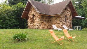 A stone building with a sloped roof, surrounded by a grassy area with two orange lounge chairs and a table with an umbrella. - Small stone house of the most unusual in the country (Habas)