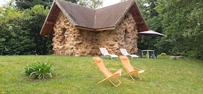 A stone building with a sloped roof, surrounded by a grassy area with two orange lounge chairs and a table with an umbrella. - Small stone house of the most unusual in the country (Habas)
