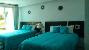 A hotel room with two beds, teal bedding, a lamp, and a view of palm trees. - Cancun Studio Front Beach (Cancún)