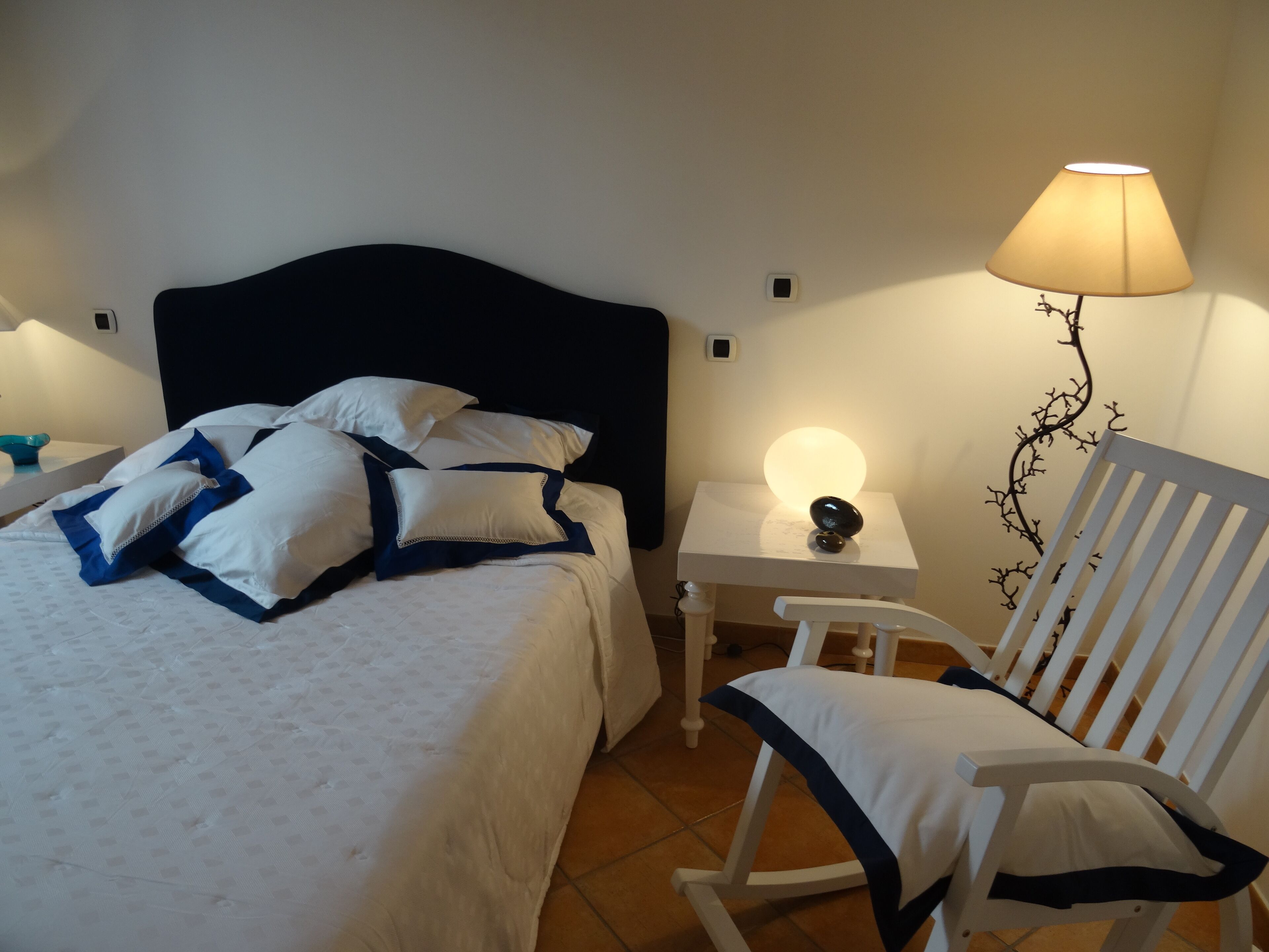 A neatly made bed with white and blue bedding, a bedside table with a lamp, and a chair with white and blue trim.