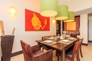 Un coin repas avec une longue table en bois, dressée pour quatre personnes, entourée de chaises en cuir marron. Trois lampes suspendues vertes sont accrochées au-dessus de la table, et un tableau abstrait rouge orne le mur à gauche.