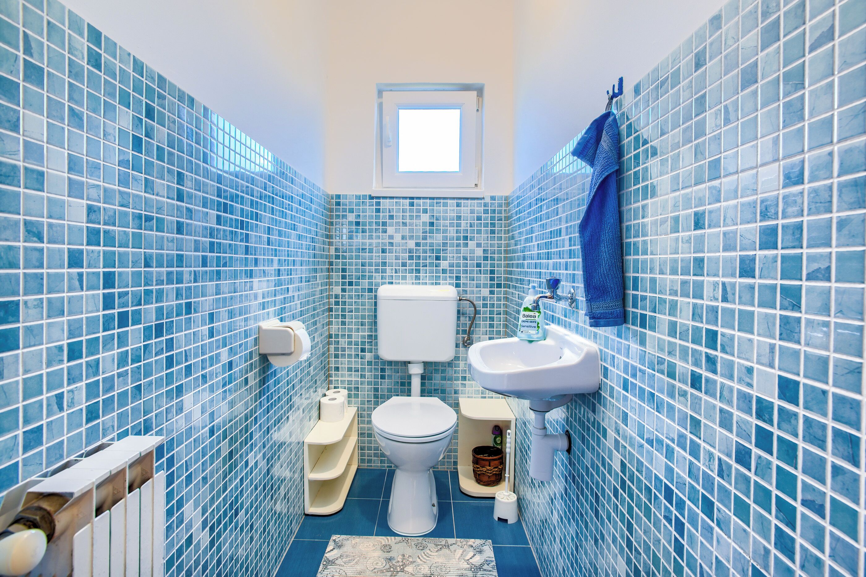 Un baño con azulejos de mosaico azules, un inodoro, un lavabo y una ventana sobre el inodoro.